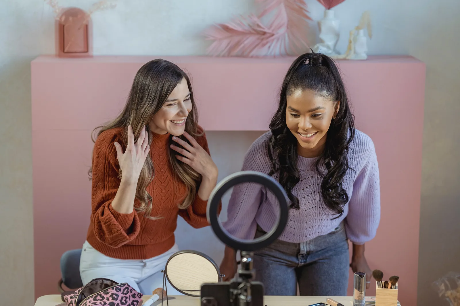 Positive multiethnic female makeup artists at table with professional supplies looking at smartphone near ring lamp while recording online lesson