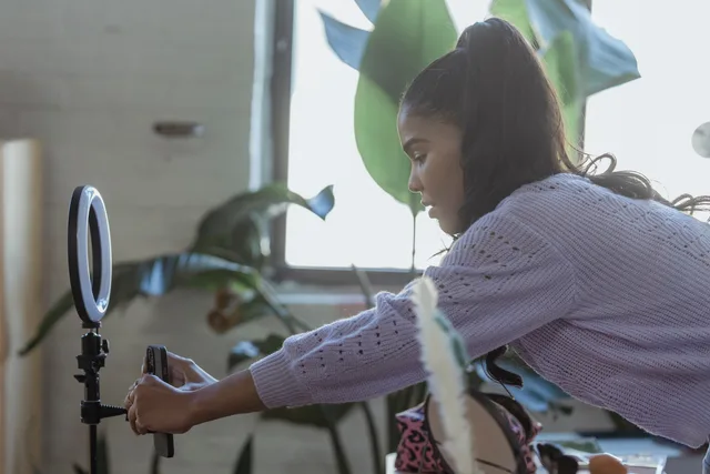 Side view of concentrated young African American female blogger with long dark hair in stylish sweater attaching smartphone to tripod with ring light before recording vlog. When to pick fixed fee, retainer or affiliate beauty creator partnerships pricing models